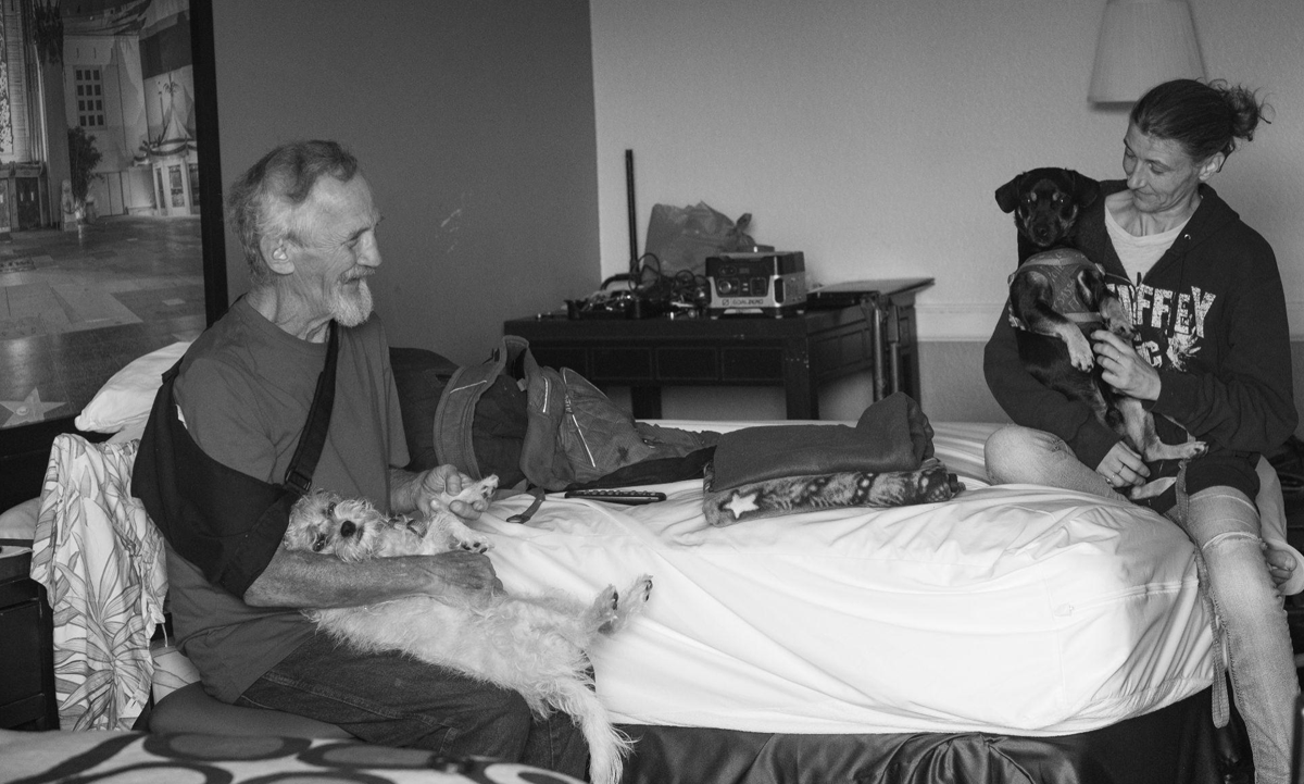 Ron Clayton and his friend Missy chat while holding their dogs in the motel where Ron lives in San Bernardino, California on June 17, 2024. Ron’s history with experiencing homelessness creates many barriers for him in accessing healthcare. He grapples with multiple health challenges that have been exacerbated from over fifteen years of living on the streets.