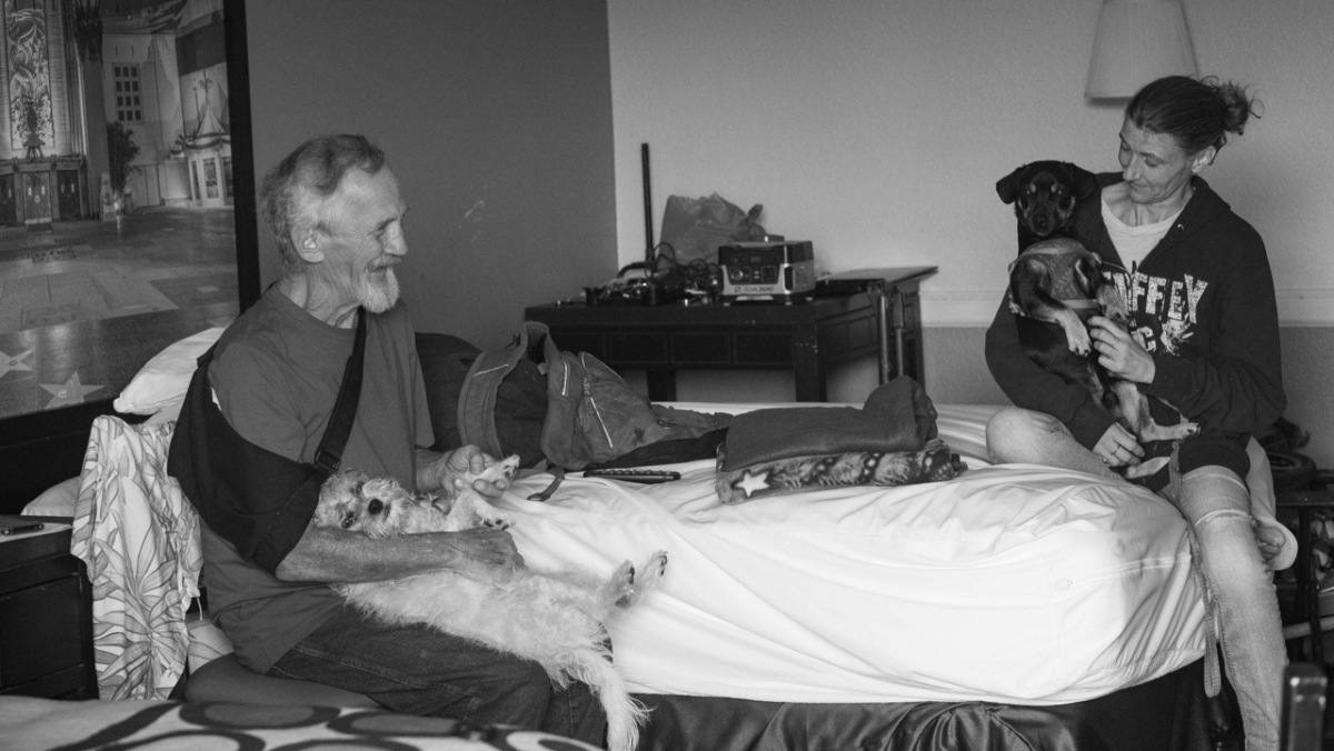 Ron Clayton and his friend Missy chat while holding their dogs in the motel where Ron lives in San Bernardino, California on June 17, 2024. Ron’s history with experiencing homelessness creates many barriers for him in accessing healthcare. He grapples with multiple health challenges that have been exacerbated from over fifteen years of living on the streets.